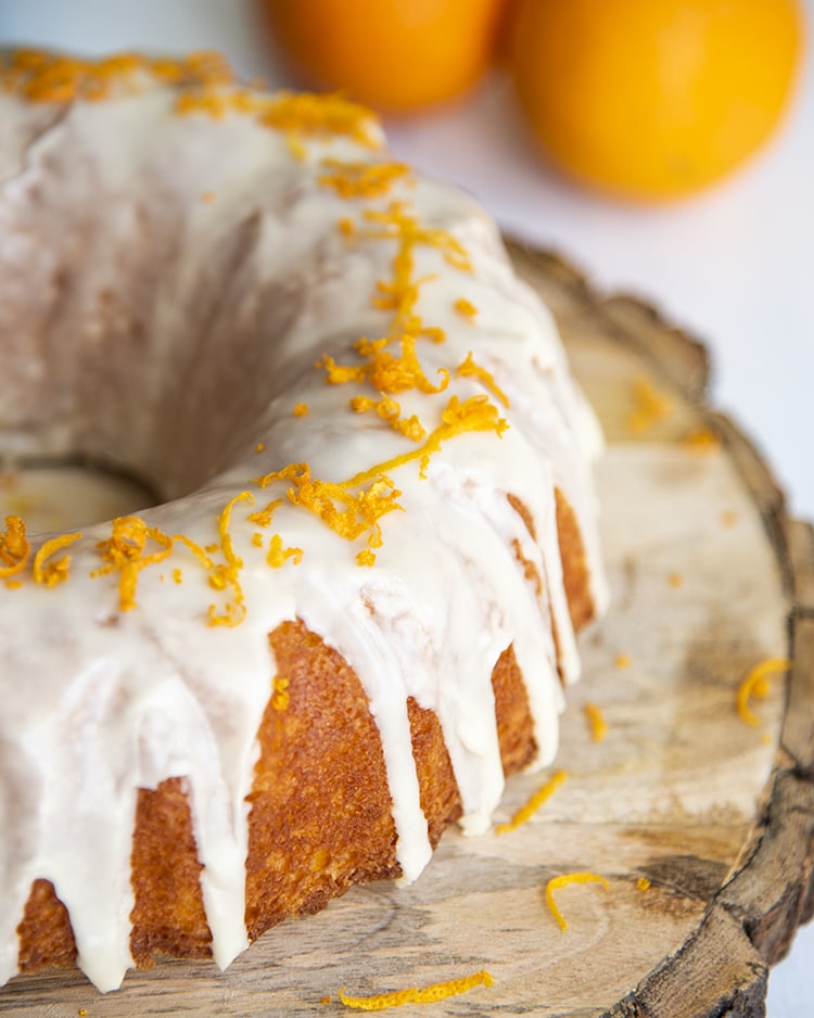A beautiful bundt cake topped with a white glaze, and sprinkled with orange zest, on a wooden tray.