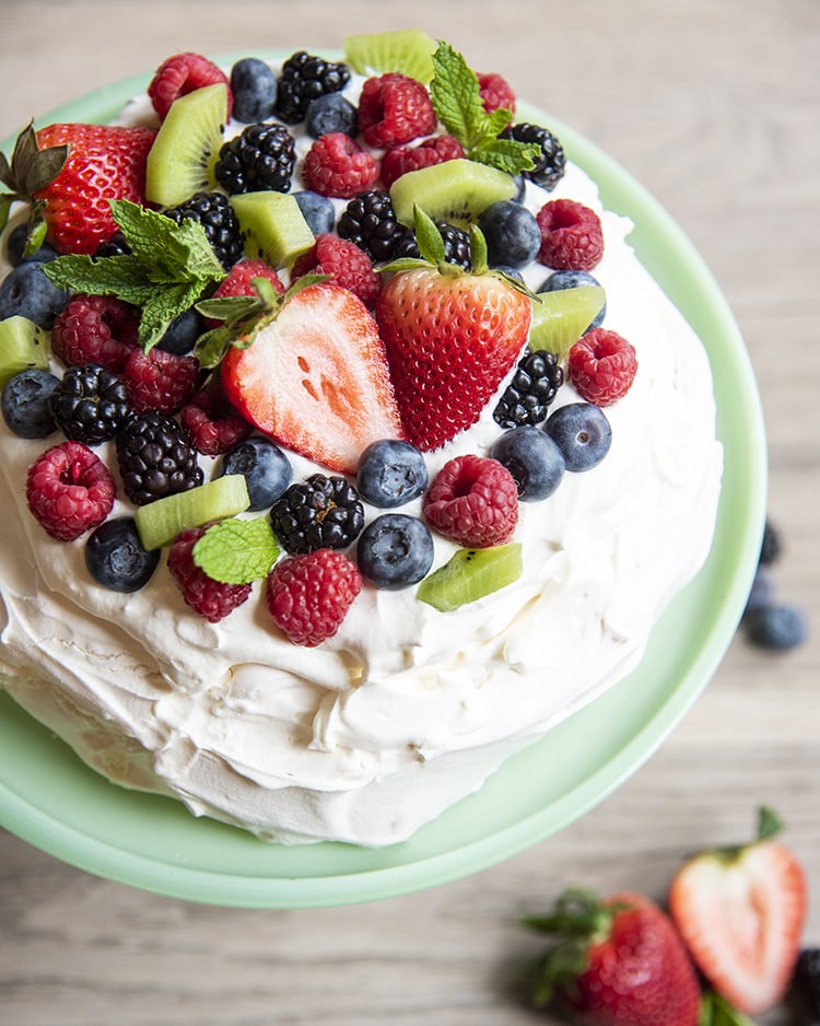 Pavlova with fruit on top sitting on top of a cake display case above image displaying the fruit.