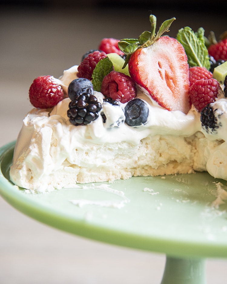 Close up of Pavlova with fruit on top sitting on top of a cake display case.