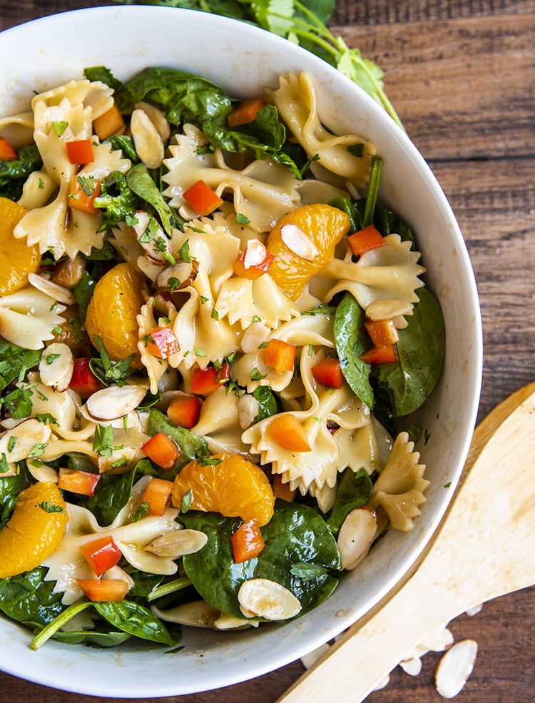 A side shot of a spinach pasta salad with bow tie noodles, mandarin oranges, spinach, cilantro, and bell peppers in a white bowl