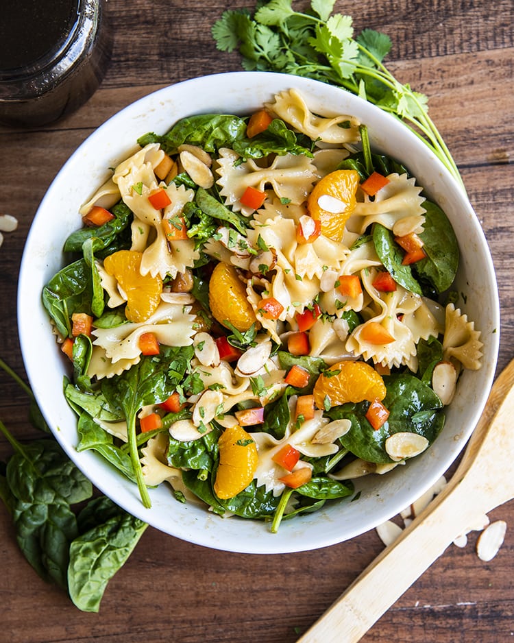An overhead shot of a spinach pasta salad with bow tie noodles, mandarin oranges, spinach, cilantro, and bell peppers in a white bowl