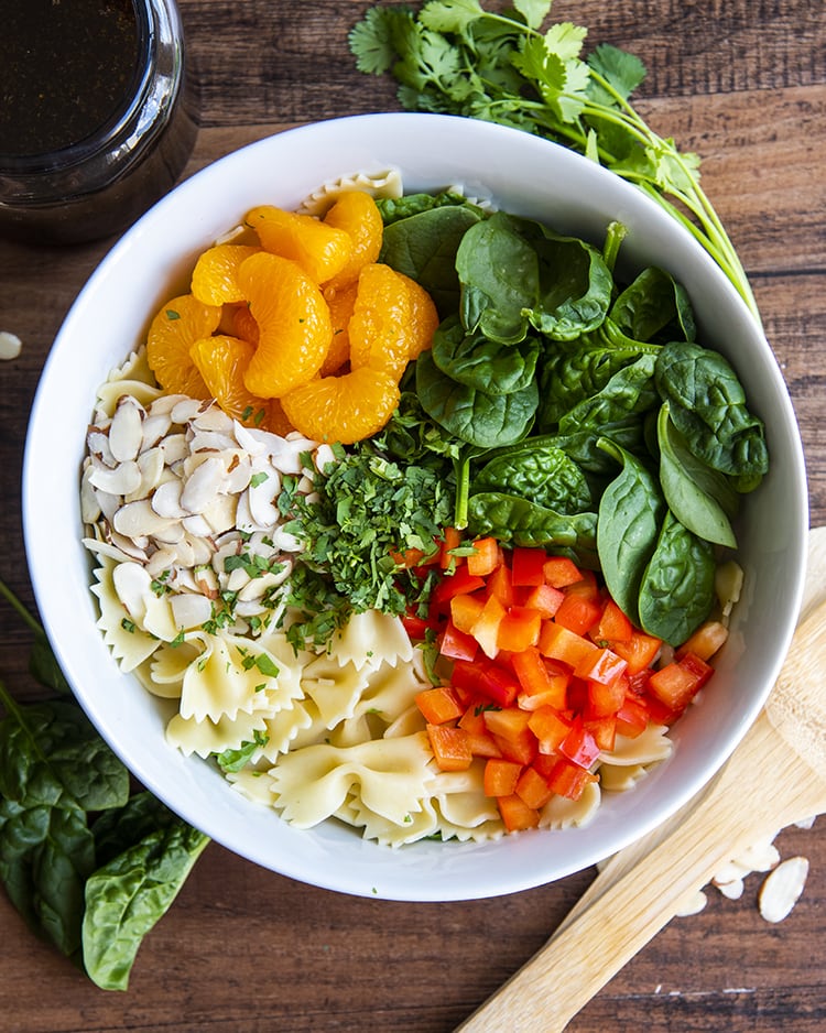 Mandarin oranges, spinach, red bell pepper, bow tie noodles, and sliced almonds in a bowl.