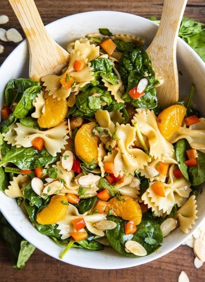 Asian pasta salad with spinach, mandarin oranges, red peppers, and sliced almonds tossed in a bowl with wooden spoons