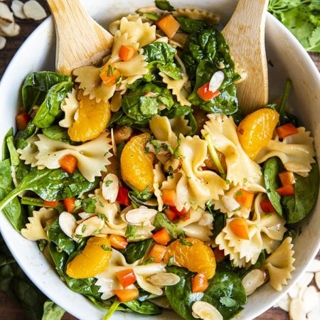 Asian pasta salad with spinach, mandarin oranges, red peppers, and sliced almonds tossed in a bowl with wooden spoons