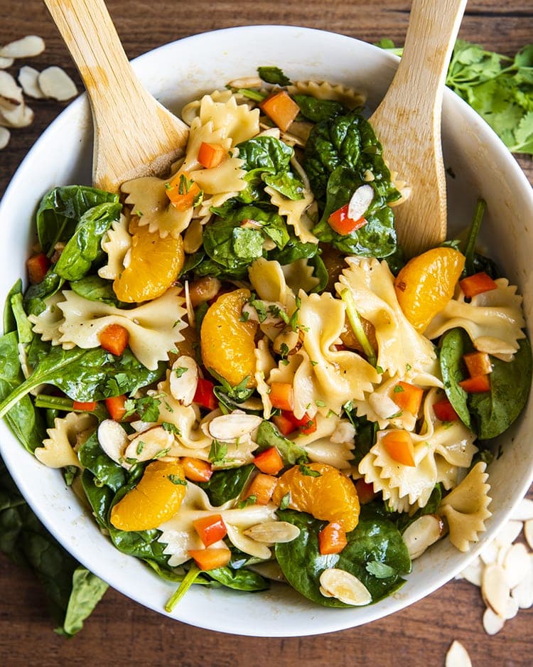 Asian pasta salad with spinach, mandarin oranges, red peppers, and sliced almonds tossed in a bowl with wooden spoons