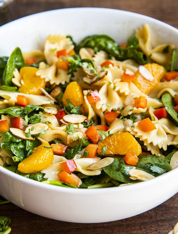 A side shot of a spinach pasta salad with bow tie noodles, mandarin oranges, spinach, cilantro, and bell peppers in a white bowl