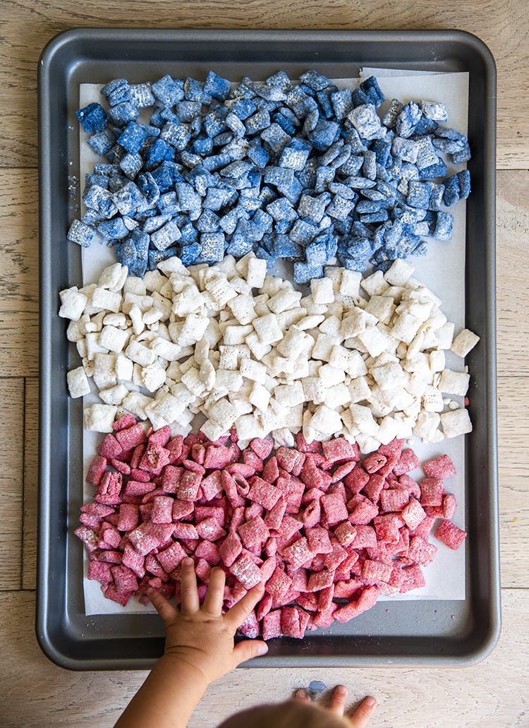 Red, white, and blue muddy buddies with m&ms and star sprinkles, separated by color on a cookie sheet with a baby hand reaching to it.