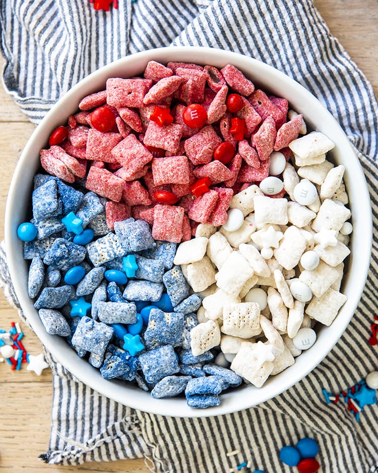 Red, white, and blue muddy buddies with m&ms and star sprinkles, separated by color in a white bowl