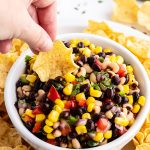 Texas caviar with a tortilla chip dipping into the bowl. The dip is full of black beans, black eyed peas, corn, bell pepper, and cilantro.