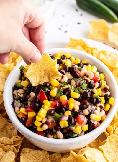 Texas caviar with a tortilla chip dipping into the bowl. The dip is full of black beans, black eyed peas, corn, bell pepper, and cilantro.