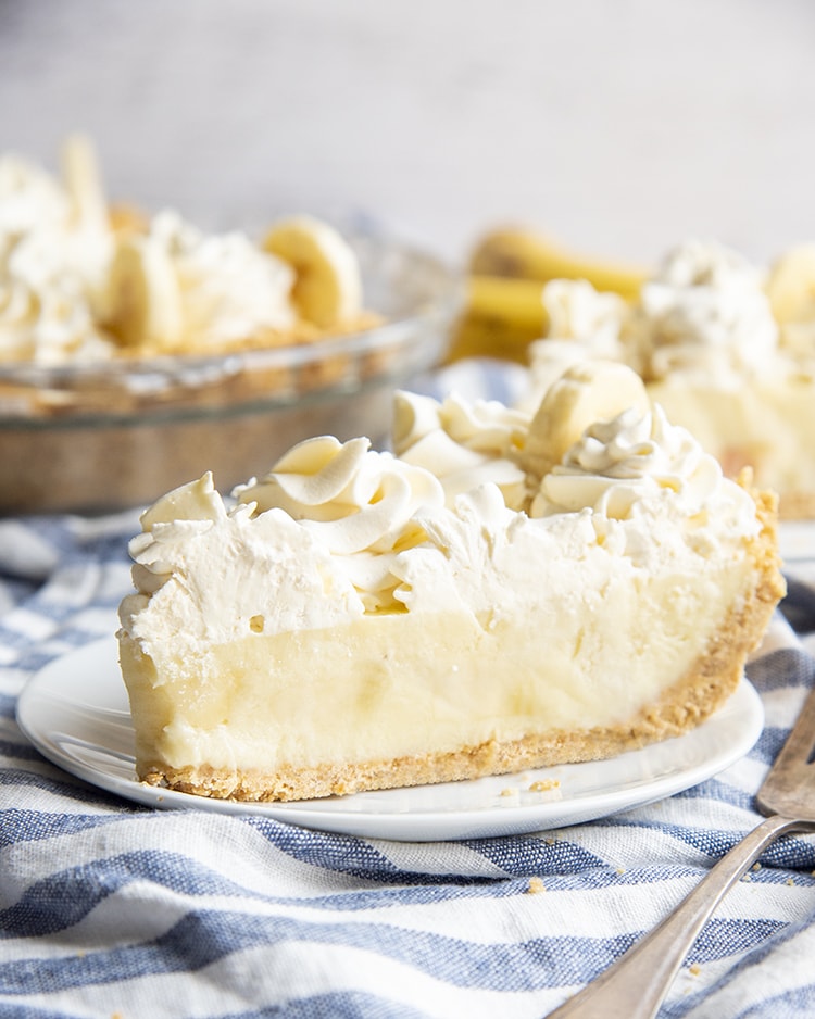 A slice of banana cream pie made from scratch. The slice is topped with whipped cream dollops and has a graham cracker crust.