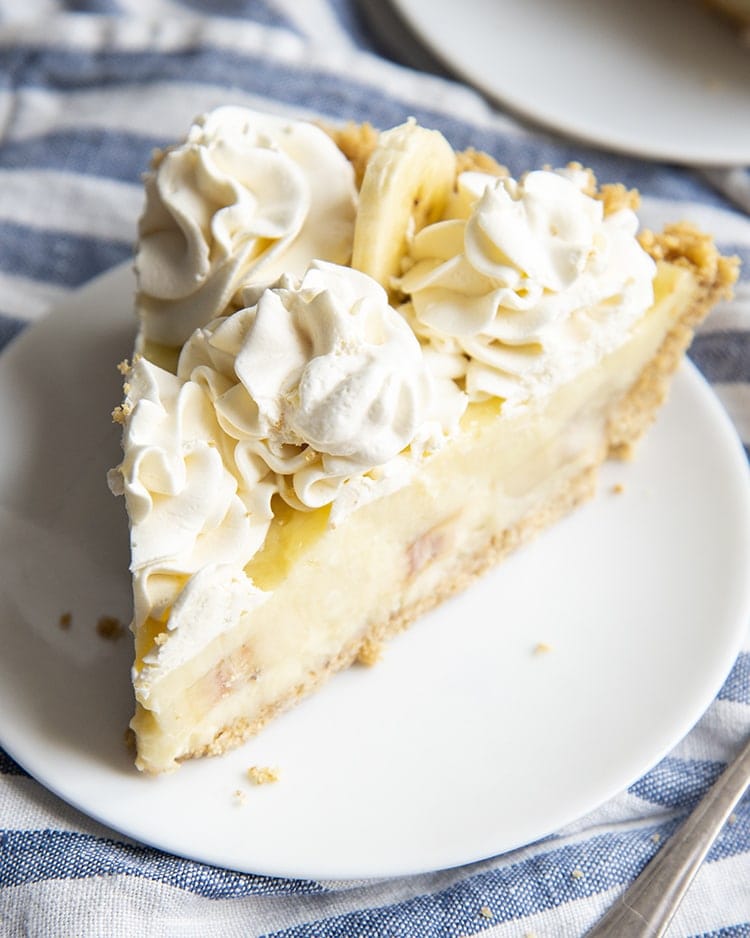 A slice of banana cream pie made from scratch. The slice is topped with whipped cream dollops and has a graham cracker crust.