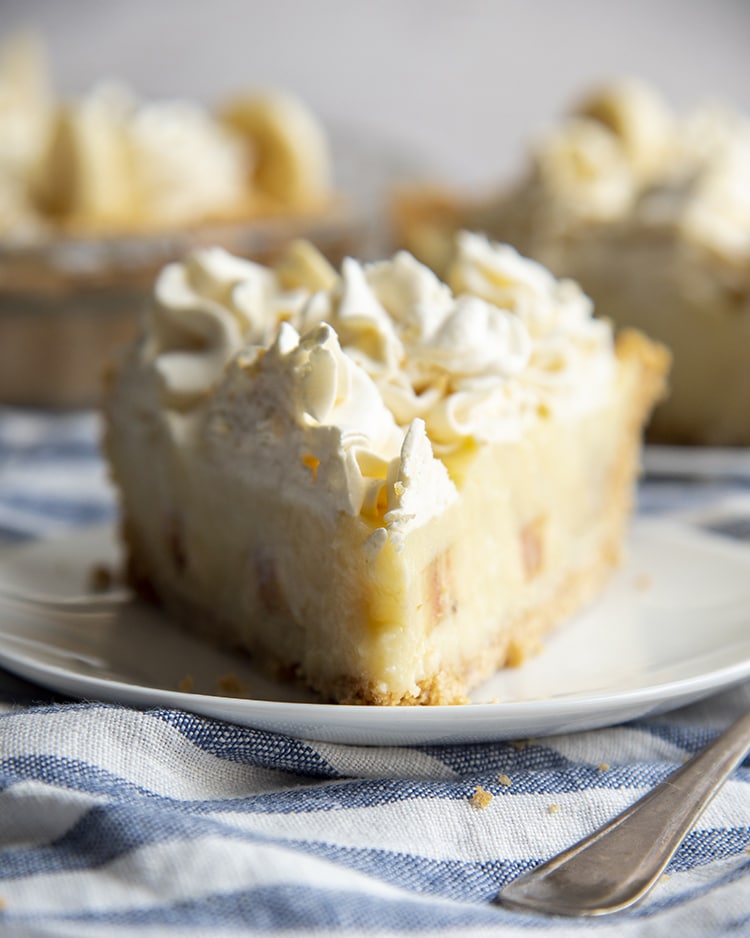 A slice of banana cream pie made from scratch. The slice is topped with whipped cream dollops and has a graham cracker crust.