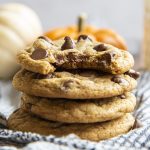 A stack of soft and chewy pumpkin chocolate chip cookies. The top cookie has a bite taken out of it.