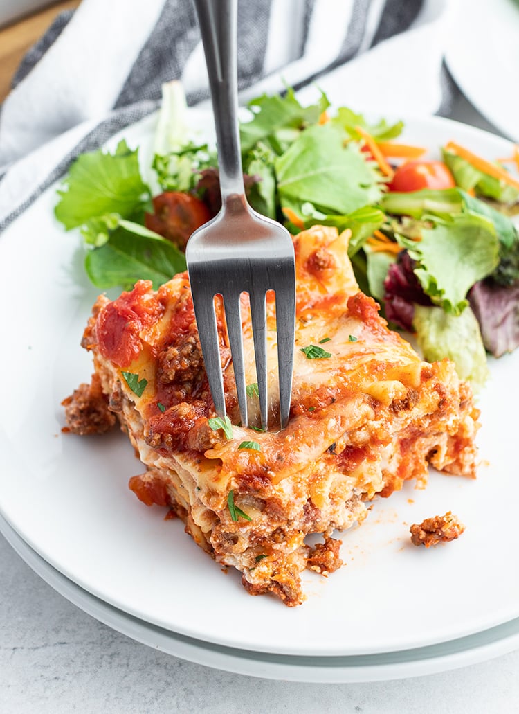 A piece of classic lasagna on a white plate with some salad.