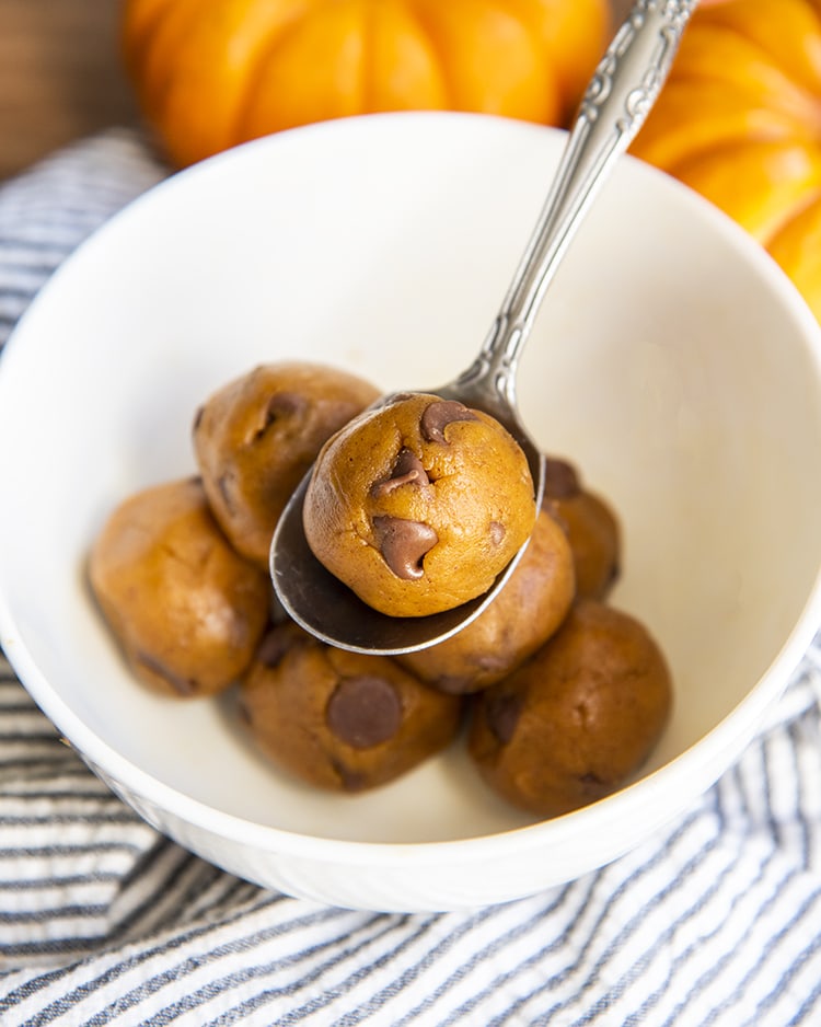 Pumpkin Cookie Dough rolled into balls in a white bowl. One cookie dough ball is on a small spoon.