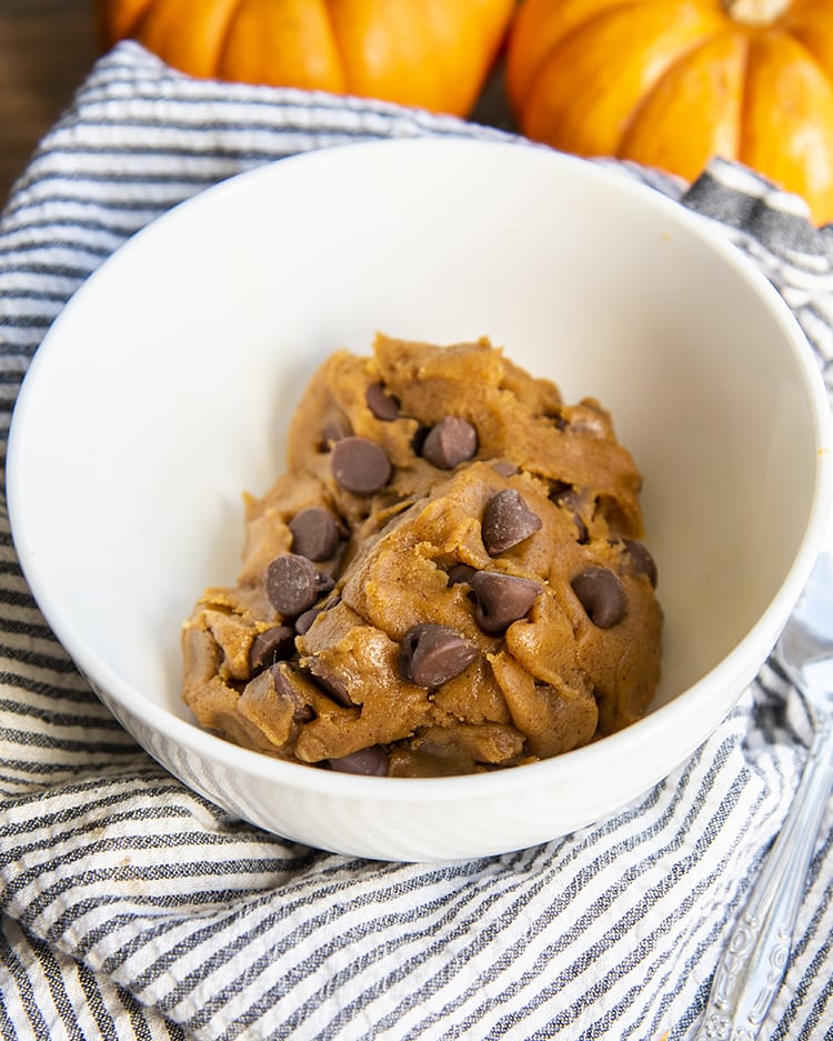Edible Pumpkin Cookie Dough with chocolate chips in a white bowl on a pinstripe linen, with small pumpkins in the back.