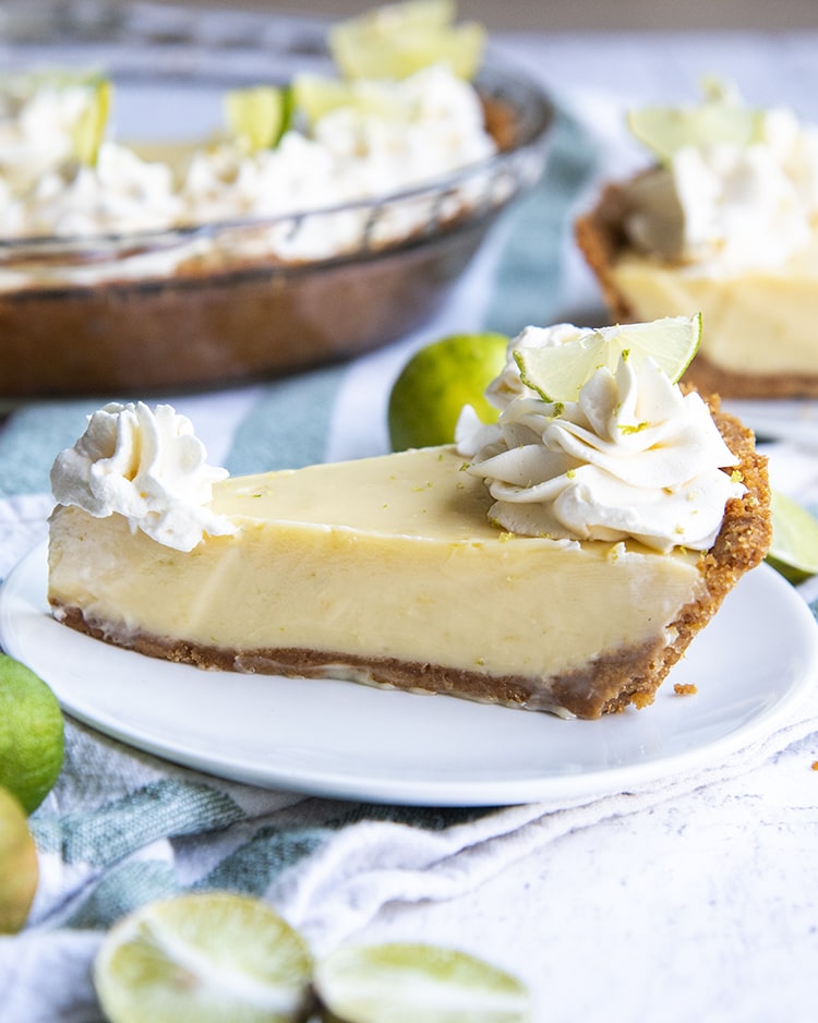 A side shot of slice of key lime pie topped with whipped cream rosettes and small lime slices, all served on a white plate.