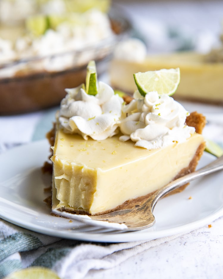 A slice of key lime pie with a bite taken out of it, opped with whipped cream rosettes and small lime slices, all served on a white plate.