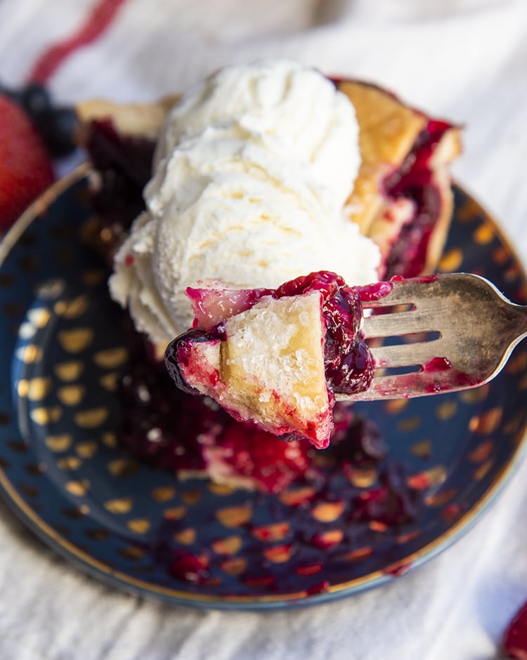 A bite of pie on a fork with the slice of berry pie on a plate behind it.