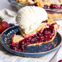 A slice of mixed berry pie on a blue plate showing all the juicy red berries in the middle with a lattice pie crust on top, with a scoop of vanilla ice cream on top.
