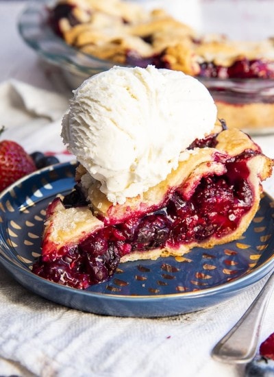 A slice of mixed berry pie on a blue plate showing all the juicy red berries in the middle with a lattice pie crust on top, with a scoop of vanilla ice cream on top.