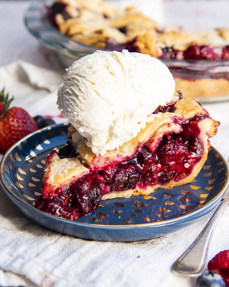 A slice of mixed berry pie on a blue plate showing all the juicy red berries in the middle with a lattice pie crust on top, with a scoop of vanilla ice cream on top.