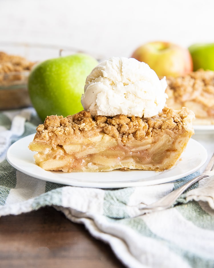 A slice of apple pie on a white plate, topped with vanilla ice cream, with apples behind it.