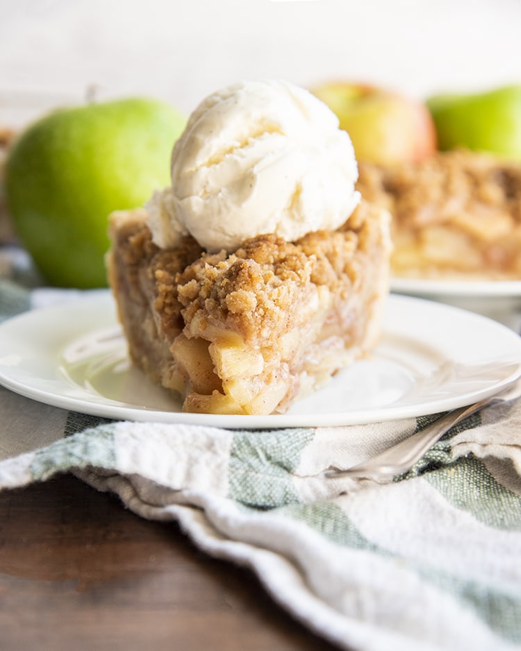 A slice of apple pie on a white plate, topped with vanilla ice cream, with apples behind it.