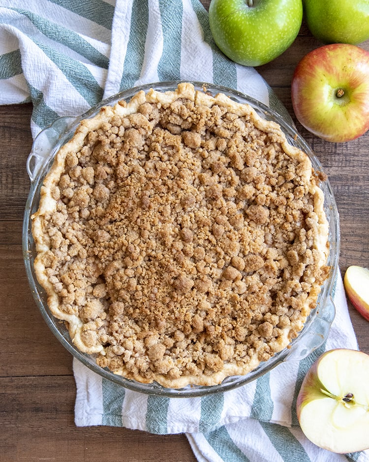A whole dutch apple pie in a pie pan, showing the streusel crumb on the top. On the side of the pie are some apples.