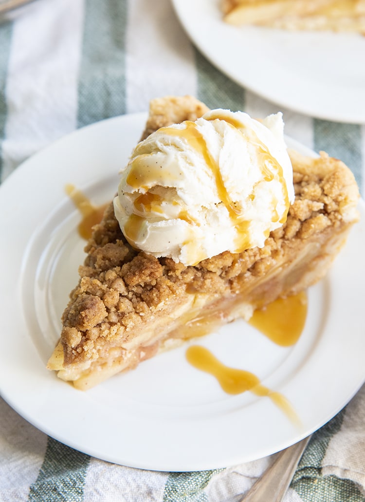 A slice of apple pie topped with a streusel crumb, and a scoop of vanilla ice cream, drizzled with caramel.