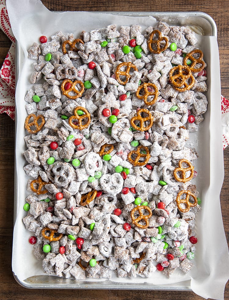 Christmas Muddy Buddies on a baking sheet lined with parchment paper. There are muddy buddies, red and green m&ms, and mini pretzels.