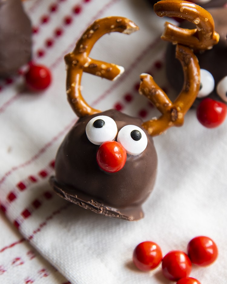 A cookie dough ball covered in chocolate, and decorated with pretzels, candy eyes and a red nose to look like a reindeer. On a white and red cloth.