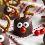 A cookie dough ball covered in chocolate, and decorated with pretzels, candy eyes and a red nose to look like a reindeer. On a white and red cloth.
