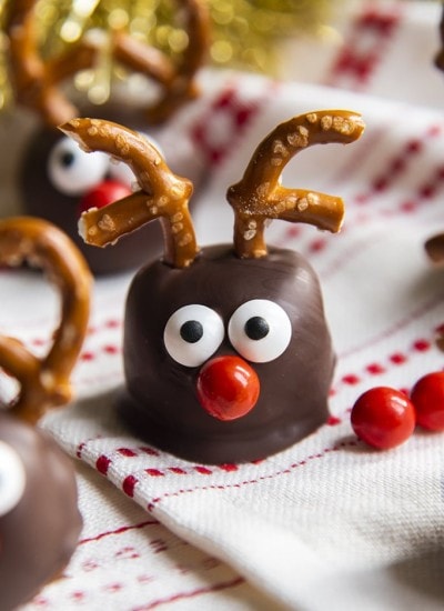 A cookie dough ball covered in chocolate, and decorated with pretzels, candy eyes and a red nose to look like a reindeer. On a white and red cloth.