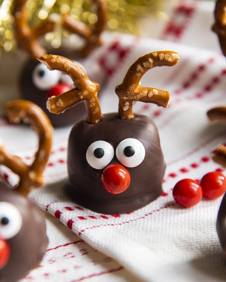 A cookie dough ball covered in chocolate, and decorated with pretzels, candy eyes and a red nose to look like a reindeer. On a white and red cloth.