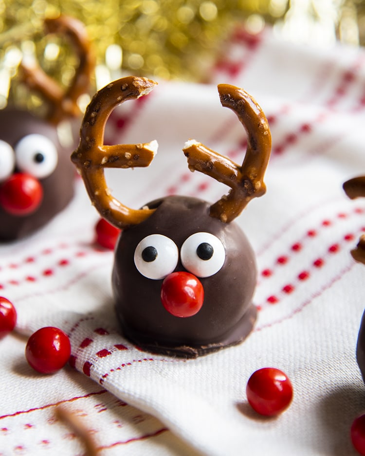 A cookie dough ball covered in chocolate, and decorated with pretzels, candy eyes and a red nose to look like a reindeer. On a white and red cloth.