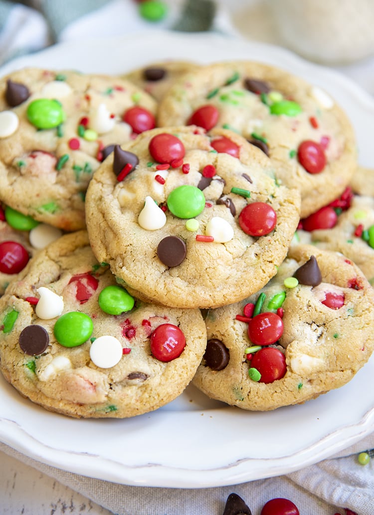 A pile of Santa's cookies on a white plate. The cookies have white chocolate chips, regular chocolate chips, green & red m&ms, and sprinkles.