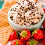 A small bowl of cannoli dip sprinkled with mini chocolate chips. The bowl is a on a plate with crepe cookies, and strawberries.