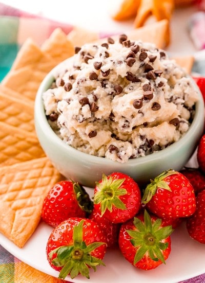 A small bowl of cannoli dip sprinkled with mini chocolate chips. The bowl is a on a plate with crepe cookies, and strawberries.