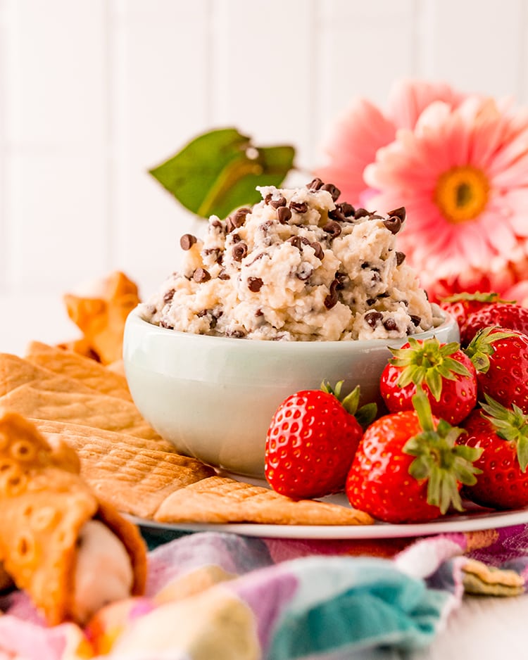 A small bowl of cannoli dip sprinkled with mini chocolate chips. The bowl is a on a plate with crepe cookies, and strawberries. There are pink flowers behind the bowl.