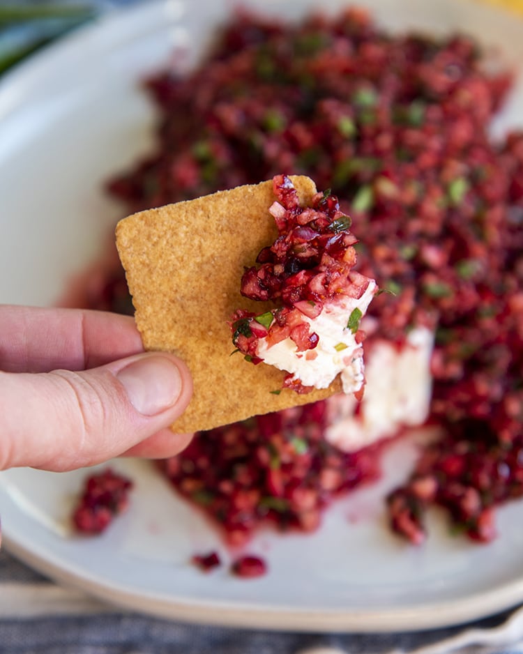 Cranberry salsa and cream cheese on a square wheat thin cracker in front of a plate of cranberry salsa over a block of cream cheese.