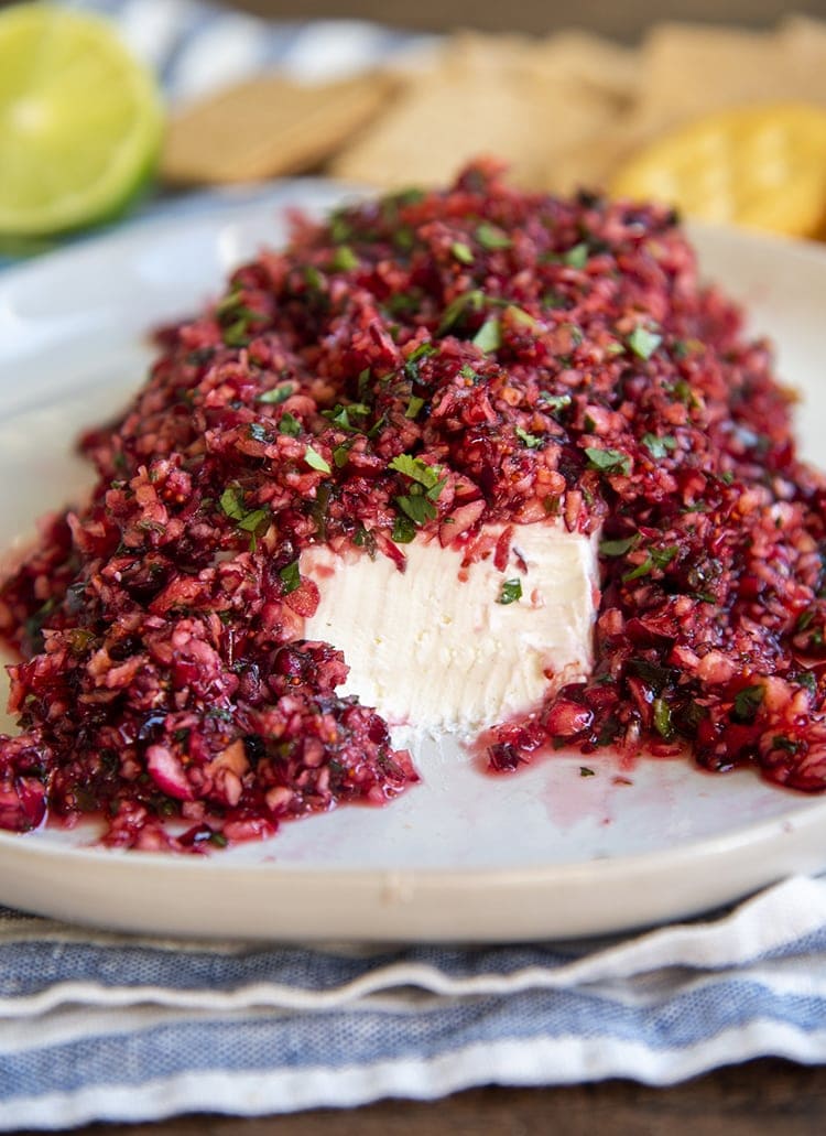 A slab of cream cheese topped with cranberry salsa, full of chopped up cranberries and cilantro. The front of the cream cheese is showing