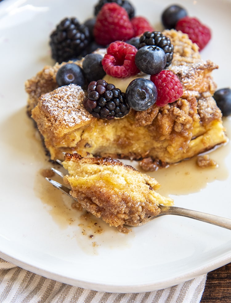 A piece of french toast casserole on a white plate, topped with fresh berries, and a fork cut a bite out of the piece.
