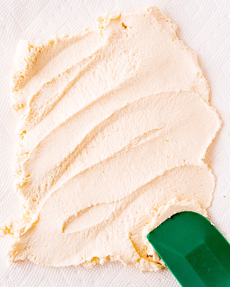 Ricotta cheese spread out on paper towels, with a green rubber spatula in the bottom right corner. 