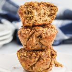 Three banana cinnamon oatmeal muffins stacked on top of each other in a tower. The top muffin is broken in half showing the inside of the muffin.