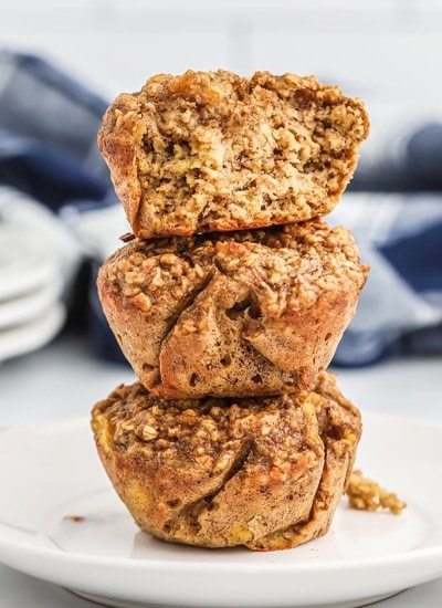 Three banana cinnamon oatmeal muffins stacked on top of each other in a tower. The top muffin is broken in half showing the inside of the muffin.