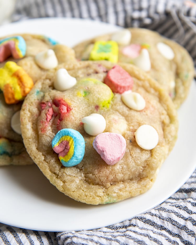 A close up of a cookie on a white plate, with two more cookies behind, the cookie is filled with white chocolate chips and lucky charms marshmallows.