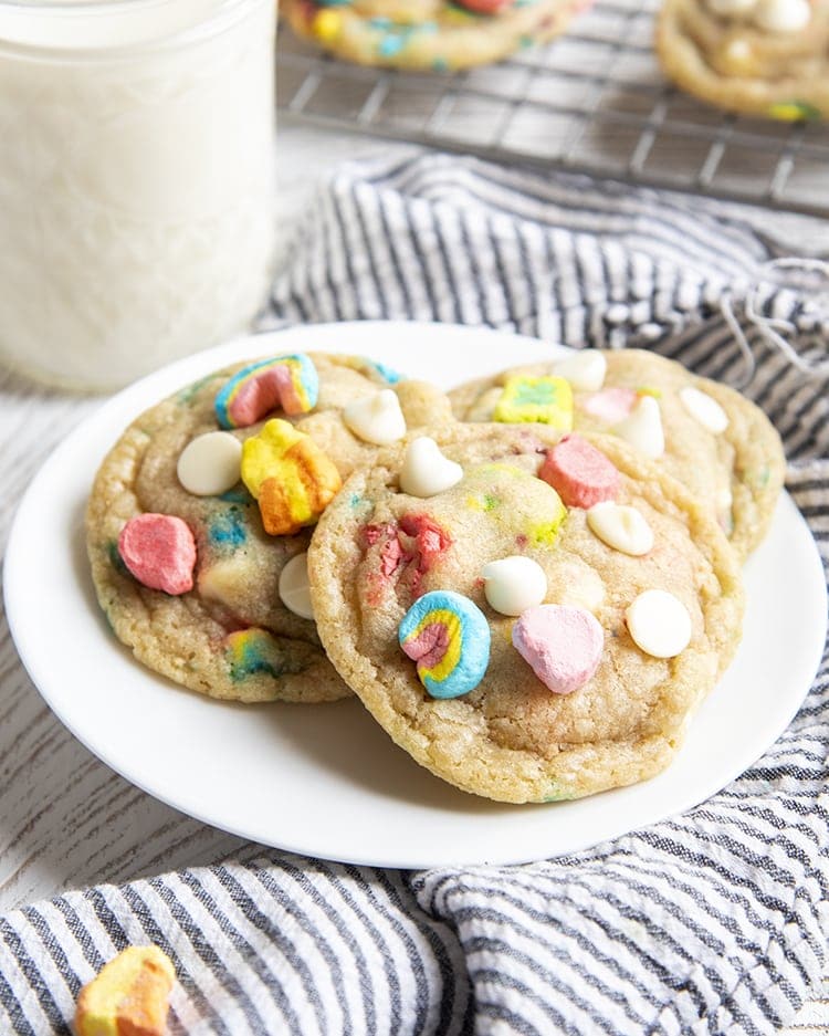 Lucky charms cookies on a small white plate with a glass of milk behind. On top of the cookies there are lucky charms marshmallows, and white chocolate chips.