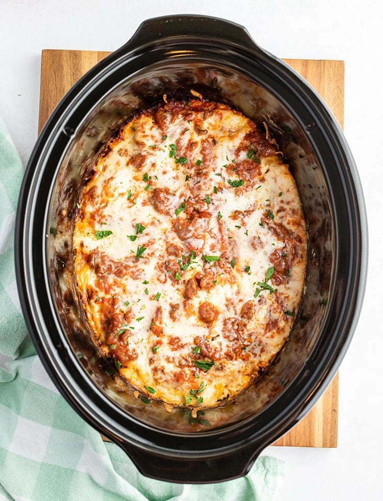 A slow cooker full of lasagna, showing cheese, and red sauce coming through, and sprinkled with parsley on top.
